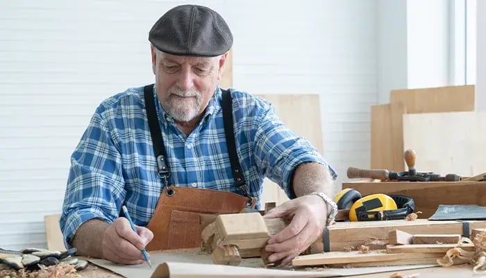 Senior man wearing a beret and apron, sketching designs on wood in a woodworking studio.