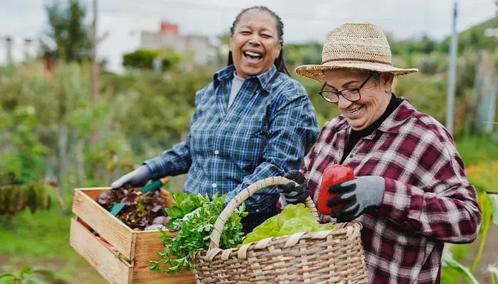 Two women laughing and holding fresh produce in a garden