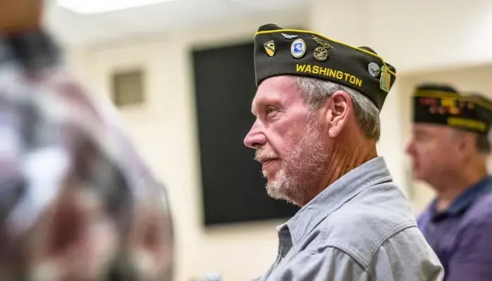 Veteran, wearing a military service cap, attentively listening during a gathering, with other veterans in the background.