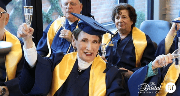 Senior resident of The Bristal wearing a mortarboard and graduation gown and sash holds up a glass to toast fellow graduates surrounded by peers