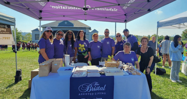 Team members of The Bristal at the Bergen-Passaic Walk to End Alzheimer's stand behind The Bristal table under a purple tent, holding yellow pinwheels to represent the impact Alzheimer's has on their lives.