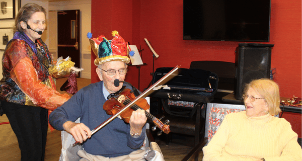 senior man playing the violin with entertainer standing behind him