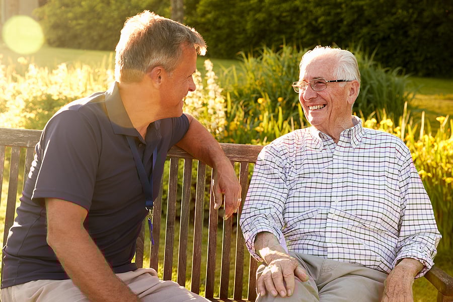 Senior male at assisted living community chats with his son on a park bench as the sun sets