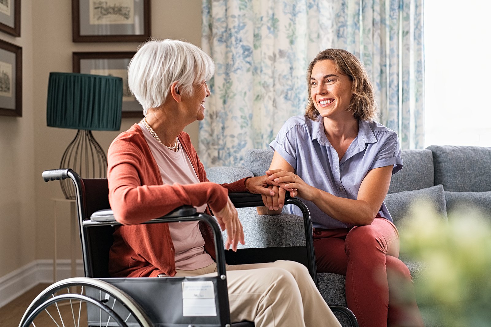 Younger woman caregiver holding hand of older woman in wheelchair in an upscale home.