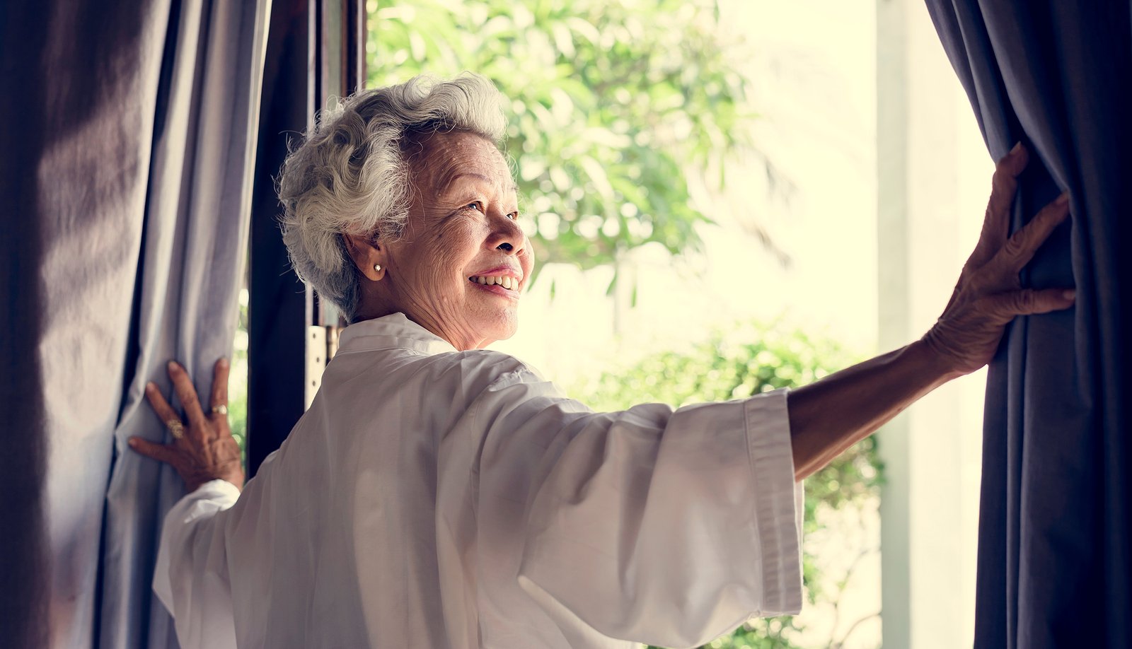 Older woman opening window curtains in her luxury senior living apartment
