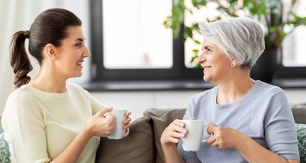 Happy smiling senior mother with adult daughter sitting on a couch, drinking tea, and talking at home.