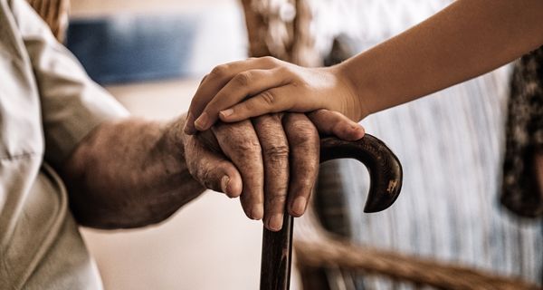 Mature man's hand resting on a cane, with a caregiver's hand resting on his hand.